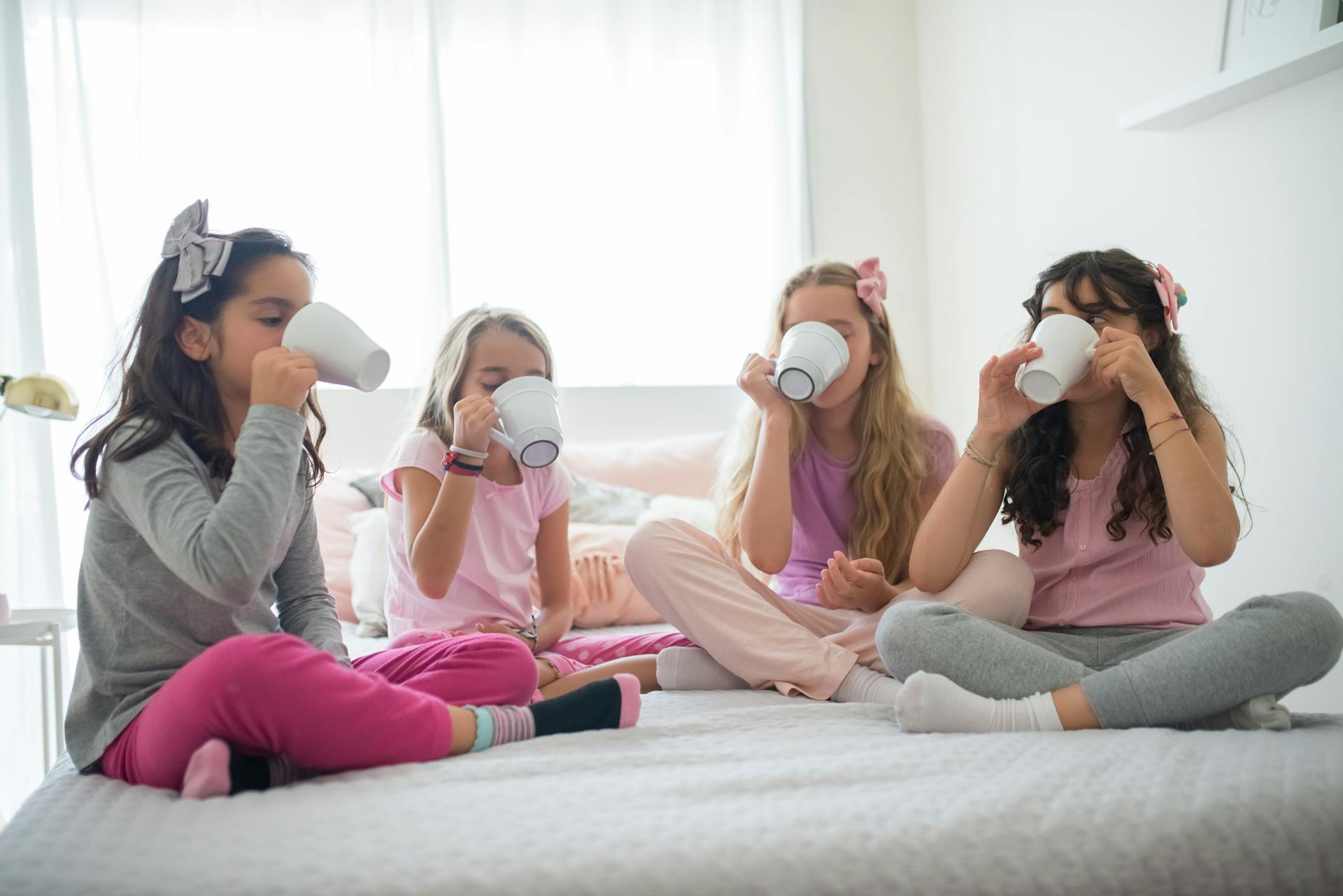 slumber party: 4 young women sitting on a bed, all of them drinking from mugs